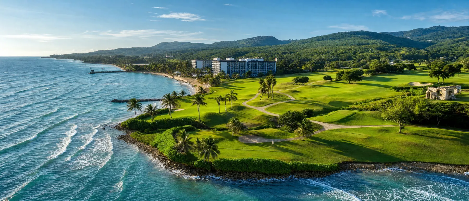 An aerial view of a coastal golf course, fairways and palms curving along a rocky shoreline with turquoise surf and green hills behind